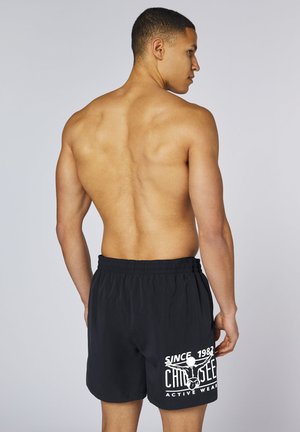 Oberkörperfreier Mann steht mit dem Rücken zur Kamera und trägt schwarze Shorts mit weißem "Since 1982 Cho Seek Active Wear" Text und Grafik am linken Bein.