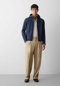 Giacca impermeabile blu navy con cappuccio, dotata di tasche frontali con zip. Abbinata a pantaloni beige a pieghe e scarpe marroni. Tessuto complessivamente liscio.