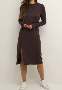 Robe marron à manches longues avec un col rond, des poignets côtelés et une fente haute sur la cuisse. Portée avec des baskets blanches, mettant en valeur une texture de tissu lisse.