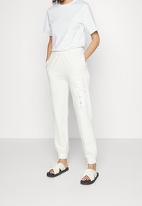 Bílý tričko v kombinaci s světle béžovými tepláky s barevným designem textu. Model nosí černé sandály s tlustými překříženými pásky.