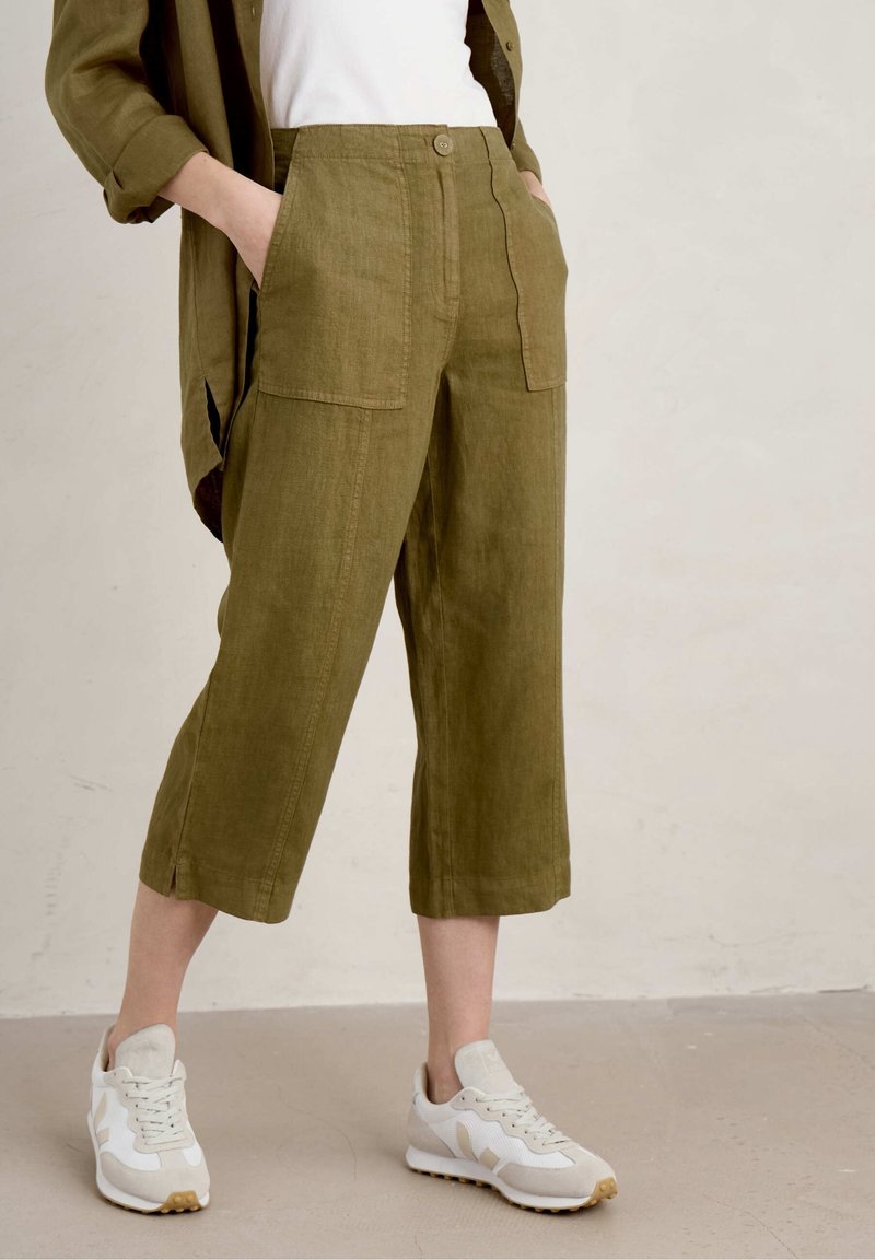 Personne portant un pantalon large court vert olive avec des poches avant, des baskets blanches, un haut blanc et une veste vert olive.