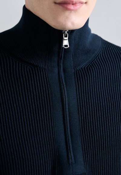 Marineblauer Strickpullover mit hohem Kragen und silbernem Reißverschluss. Verfügt über vertikale Rippungen und eine glatte Textur über der Brust.