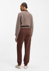 Sweater i ribstrikket brunt stof med rund halsudskæring og lange ærmer, kombineret med brune joggere med elastisk talje og lyse sko.