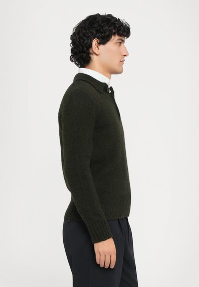 Dunkelgrüner Pullover mit strukturiertem Strick, mit einem kragenlosen Ausschnitt und gerippten Bündchen, kombiniert mit dunklen Hosen, im Profil gezeigt.