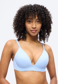 Γυναίκα με σγουρά μαλλιά που φοράει ανοιχτό μπλε σουτιέν, κοιτάζοντας μπροστά με ουδέτερη έκφραση σε απλό λευκό φόντο.