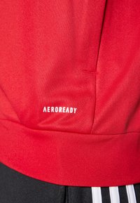Crvena sportska jakna s teksturiranim završetkom, elastičnom manžetom i bijelim logotipom "AEROREADY". Dodatne pojedinosti uključuju crni pojas.