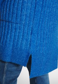 Pull en maille bleu avec texture côtelée, doté de fentes sur les côtés. Le matériau semble doux et mélangé, accentué par une teinte bleu profonde.