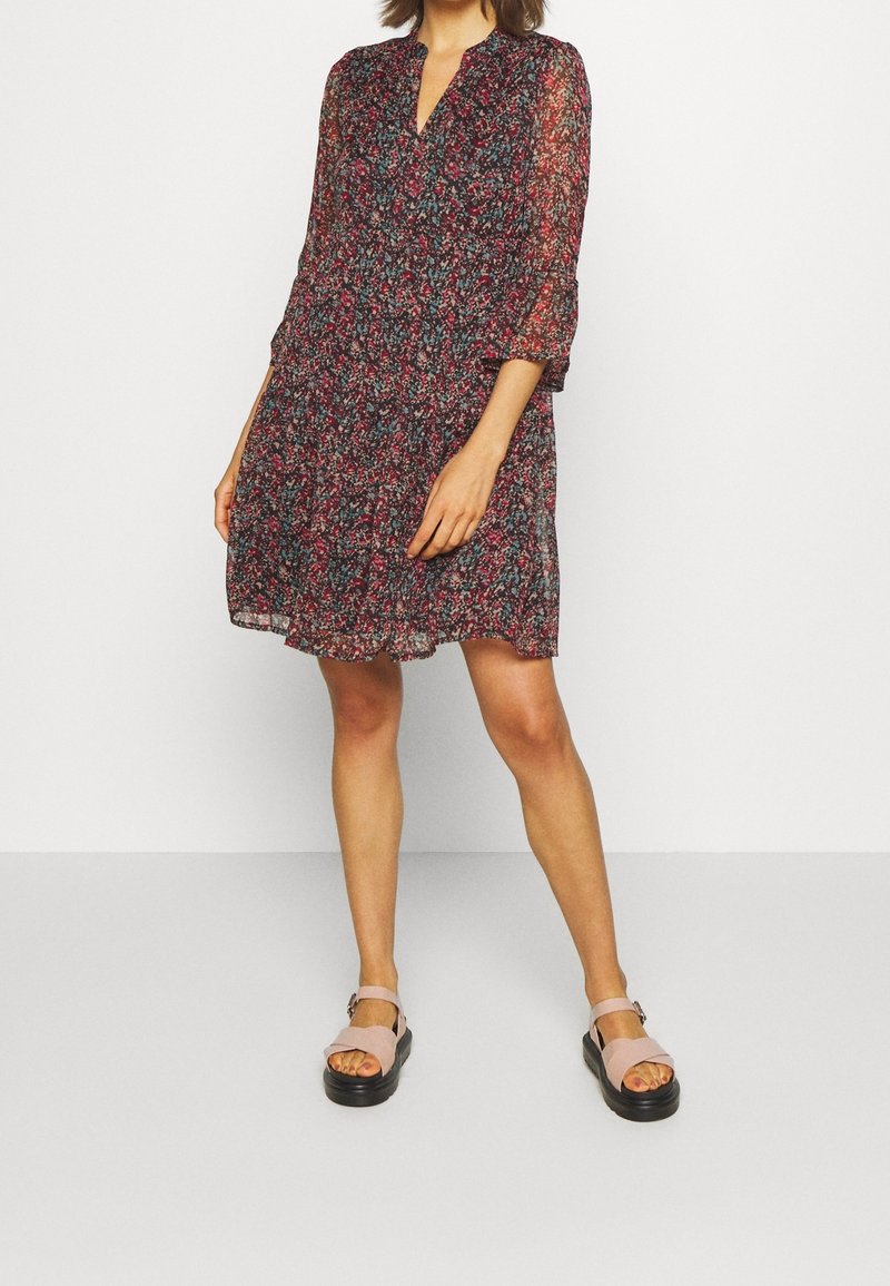 Femme portant une robe fleurie longue jusqu'aux genoux, manches longues, noire, rouge et bleue, avec des sandales compensées rose clair, debout sur un fond blanc.