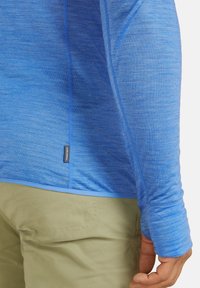 Camisa de manga larga en tejido azul degradado, con patrón texturizado, costuras planas y una etiqueta en el lateral. Combinada con pantalones beige.