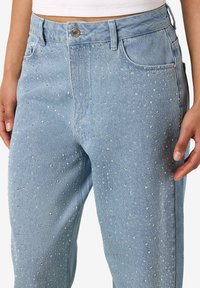 Šviesiai mėlynos, aukštu liemeniu džinsinės kelnės su sidabriniais blizgučiais, priekyje sagomis užsegamos ir su kišenėmis, dėvimos su balta trumpa palaidine.