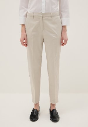 Person trägt beige maßgeschneiderte Hose, weißes Hemd mit Knöpfen und schwarze Loafers, steht vor einem schlichten hellen Hintergrund.