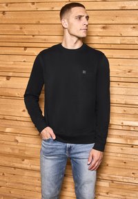 Schwarzes Crewneck-Sweatshirt aus glattem, weichem Material mit einem kleinen Logo auf der Brust. Kombiniert mit hellblauen Jeans vor einem Holz-Hintergrund.