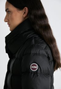Zwarte pufferjack met een hoge kraag, voorzien van een ronde paarse logo patch met het label "COLMAR" op de linker mouw. Zachte, glanzende textuur.
