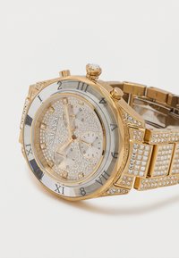 Ρολόι Swarovski σε χρυσό τόνο με πρόσωπο και μπάντα διακοσμημένα με κρύσταλλα, ενδείξεις ώρας με λατινικούς αριθμούς και τρεις μικρούς χρονογράφους.