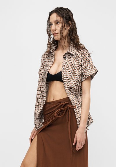 Νεαρή γυναίκα με βρεγμένα μαλλιά φοράει ανοιχτό πουκάμισο με σχέδια και κοντά μανίκια πάνω από μαύρο σουτιέν και καφέ φούστα τύπου σάκου με άνοιγμα στο πλάι.