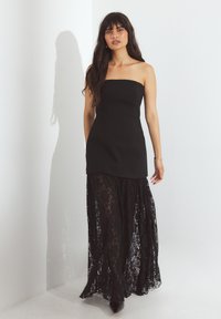 Moteris su ilgais tamsiais plaukais, vilkinti juodą suknelę be petnešėlių su nėriniuotu permatomu sijonu, stovi prieš baltą sieną su šešėliu.