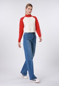 Κόκκινο και κρεμ πουλόβερ με φερμουάρ και μακριά μανίκια, σε συνδυασμό με μπλε τζιν ίσιας γραμμής και λευκά αθλητικά παπούτσια με μαύρες ρίγες.