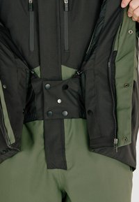 Schwarz und grüne Jacke aus glattem Stoff, mit doppelt Reißverschlüssen, Druckknopfverschlüssen und einem elastischen Bund. Verfügt über große Innentaschen für Stauraum.