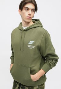 Olīvu zaļa Nike kapuce ar priekšējo kangaru kabatu, kurā redzams balts teksts "TIKAI AUTORIZĒTĀM PERSONĀM" un logo kreisajā krūtī.