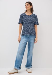Camiseta de manga corta de color marino con un estampado de hojas. Tejido suave, corte relajado. Combinada con unos vaqueros de corte suelto de color azul claro y zapatillas blancas.