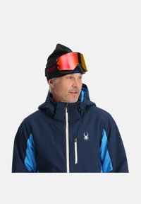 Marineblaue Skijacke mit weißem Reißverschluss und Spinnenlogo. Schwarze Mütze und verspiegelte Skibrille mit gelben und roten Akzenten.