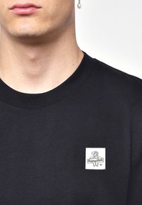 Mustan puuvillapaitasi, jossa on pyöreä kaula-aukko ja lyhyet hihat. Edessä on neliömäinen valkoinen kangastagi, jossa on sarjakuvamaalaus.