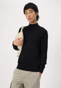 Maglione nero a collo alto con vestibilità slim, polsini a coste e un piccolo logo. Il modello tiene una borsa a tracolla beige. Indossa pantaloni cargo grigi chiari.