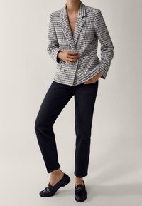 Personne portant un pantalon droit noir, des mocassins noirs avec un détail doré, et un blazer à carreaux noir et blanc avec des boutons dorés.