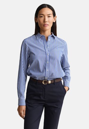 Woman wearing a blue and white vertical striped button-up shirt tucked into navy pants with a brown belt, standing with one hand in pocket.