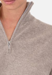 Pull beige à col zippé en tissu tricoté doux. Il comporte une fermeture éclair argentée et une coupe décontractée avec une texture côtelée au col et à l'ourlet.