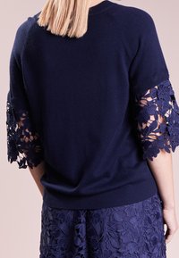 Mujer vestida con una blusa de manga corta azul marino con recortes de encaje floral y una falda de encaje floral a juego, mostrada desde la espalda.