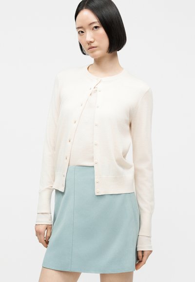 Γυναίκα με ίσιο μαύρο καρέ χτένισμα που φοράει ανοιχτό μπεζ πουλόβερ με κουμπιά και ανοιχτό μπλε φούστα σε λευκό φόντο.