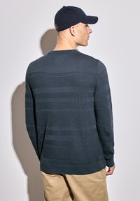Navy blue knit sweater with horizontal stripe patterns. Long sleeves and crew neck. Worn with khaki pants and a navy cap. Side view.