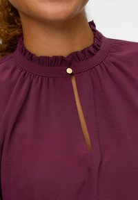 Blusa bordeaux con colletto arricciato, apertura a chiave fermata da un bottone dorato e una texture di tessuto liscia.