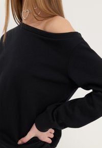 Sweat-shirt noir à épaules dénudées avec manches longues, col et poignets côtelés, texture lisse, coupe ajustée et détails sur les coutures latérales.