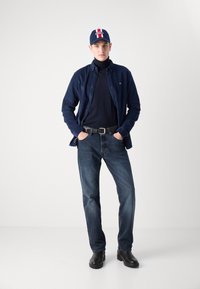 Camicia blu con bottoni sopra un dolcevita nero, abbinata a jeans scuri e scarpe nere. Un cappellino con strisce rosse e bianche completa il look.