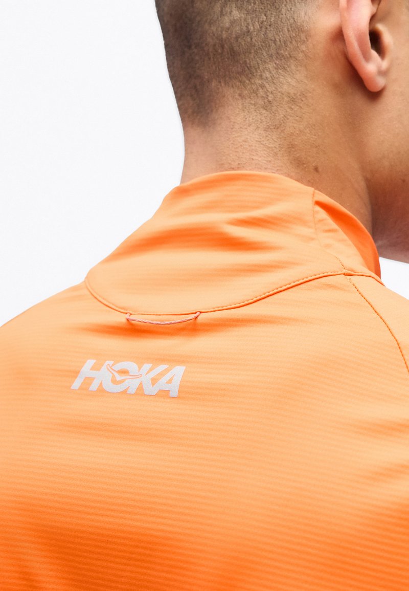 Nahaufnahme des Nackens und der Schulter einer Person, die ein oranges HOKA-Sportshirt trägt, mit sichtbarem Markenlogo im oberen Rückenbereich.
