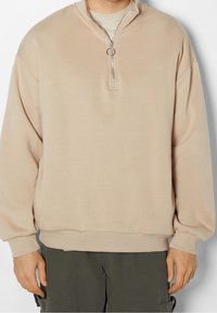 Sudadera beige con cremallera y cuello alto, fabricada en una tela suave. Presenta puños y dobladillo acanalados, un corte relajado y una cremallera plateada.