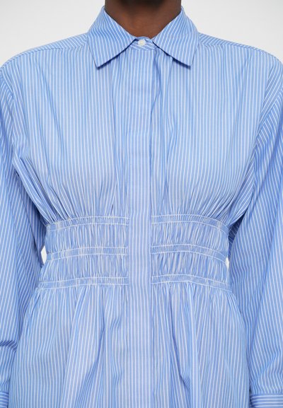 Camicia a righe azzurro chiaro con colletto, dotata di arricciature elastiche in vita e maniche lunghe. Tessuto dalla consistenza liscia.