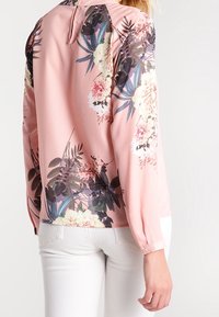 Blusa floreale rosa con maniche lunghe, caratterizzata da un dettagliato motivo floreale in tonalità di bianco, verde e nero. Tessuto morbido e polsini con bottoni.