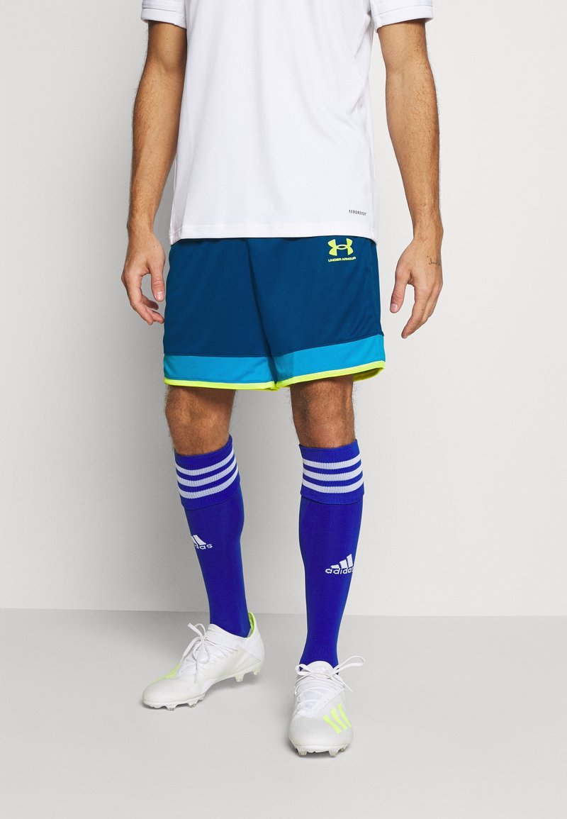 Homme portant une chemise blanche, un short bleu Under Armour avec une bordure bleu clair et néon, des chaussettes hautes Adidas bleues et des crampons blancs avec des détails néon.