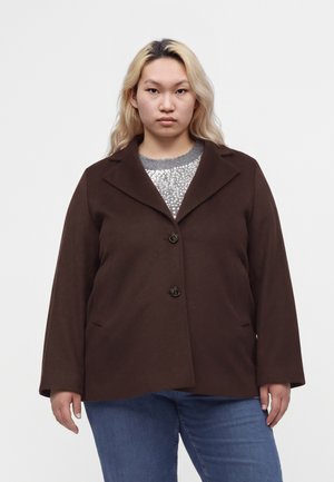 Femme aux longs cheveux blonds portant un manteau boutonné marron foncé par-dessus un haut gris à motifs et un jean bleu, debout devant un fond uni.