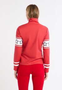 Czerwony top z długim rękawem z wysokim kołnierzem i paskami na rękawach z czarnymi wzorami gwiazd. Miękki materiał o dopasowanym kroju.