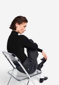 Schwarzer gerippter Rollkragenpullover mit eng anliegenden Ärmeln, getragen mit grauen gestreiften Hosen und schwarzen Schuhen, sitzt auf einem transparenten Plastikstuhl.