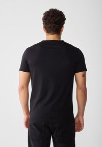 Svart t-skjorte med korte ermer laget av bomull, med rund hals. Synlig tatovering på armen. Brukt med mørke denimjeans.