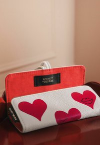Portefeuille en cuir blanc avec des graphiques de cœur rouge et de lèvres, doté d'une finition texturée et d'un accent en suède rouge contrastant, ainsi qu'une étiquette de marque.