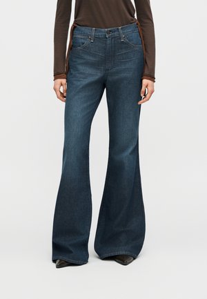 Γυναίκα που φοράει σκούρο μπλε τζιν με ψηλή μέση και φαρδιά παντελόνια και ένα καφέ μακρυμάνικο τοπ, στέκεται μπροστά σε ένα απλό λευκό φόντο.