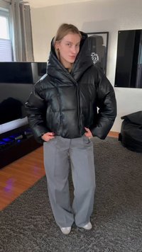 Személy fekete, túlméretezett fazonú pufi dzsekit és szürke, bő szárú nadrágot visel, szőnyeggel borított padlón áll egy nappaliban.