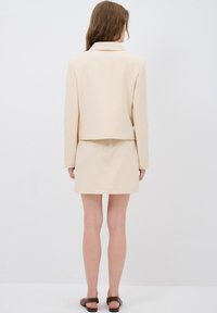 Beige blazer en bijpassende rok. Gemaakt van een gestructureerde stof, met een notch-kraag en drie knopen op de mouwen. Bruine platte sandalen.