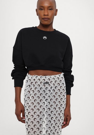Black cropped sweatshirt with a round white logo on the chest, paired with sheer white pants featuring a circular brown pattern.